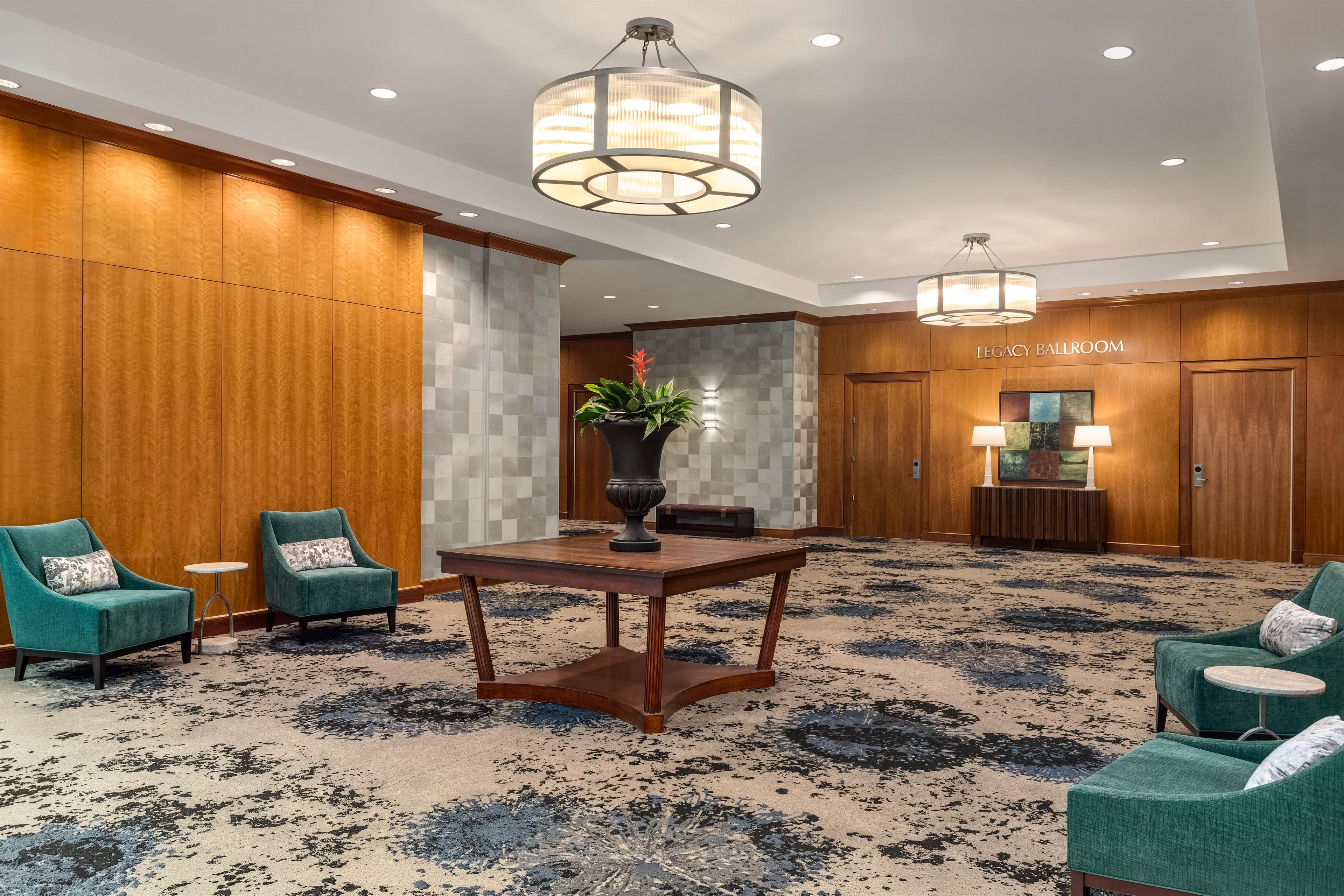 A hotel lobby with a large chandelier, green armchairs, a round table, and a 'Legacy Ballroom' sign.