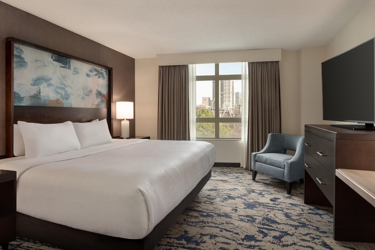 Hotelkamer met een groot bed, een bureau, een stoel, een televisie en een raam met uitzicht op gebouwen en een reuzenrad.