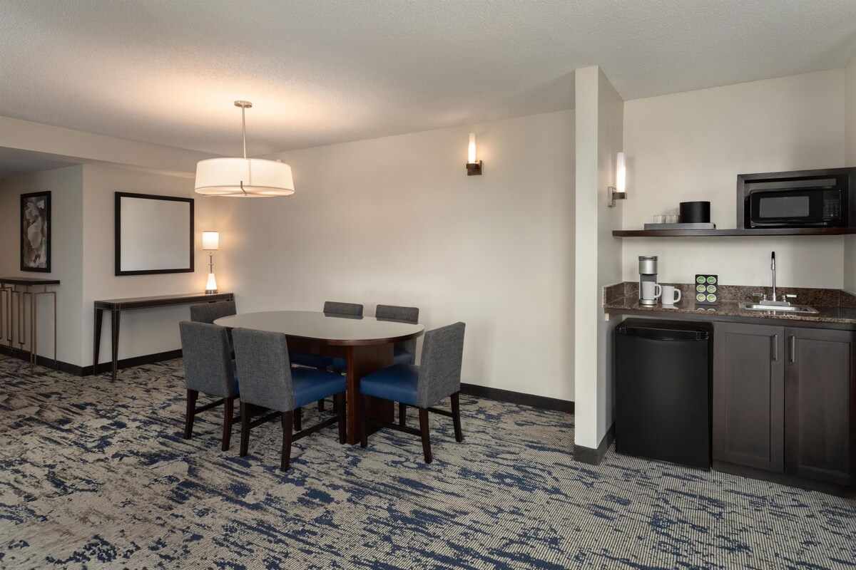 Een hotelkamer met een eettafel, stoelen, een keukenhoek met een magnetron en een koffiezetapparaat.
