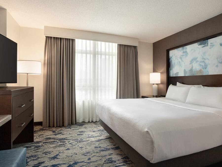 A hotel room with a bed, a TV, a desk, a lamp, and a painting on the wall.