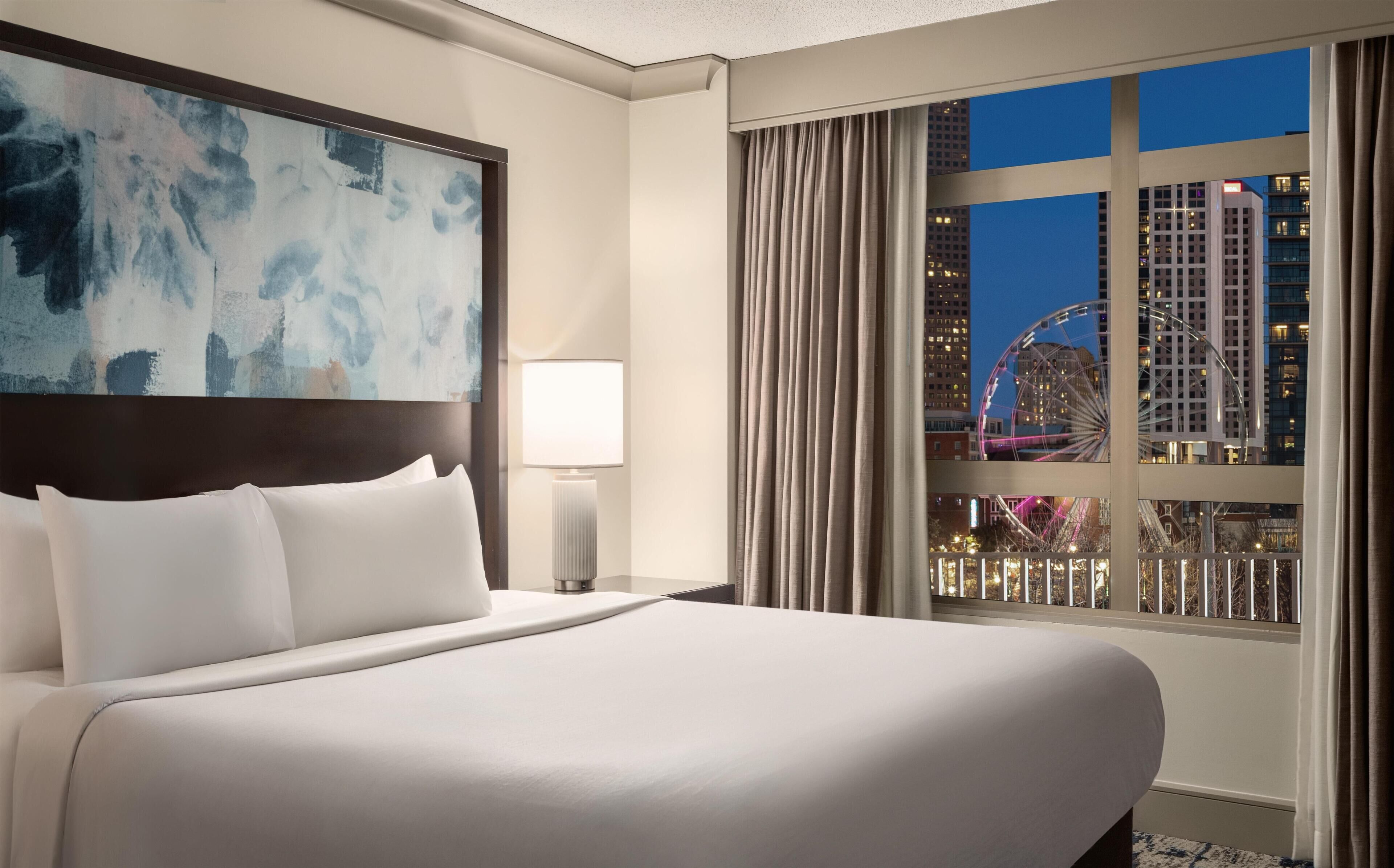 A hotel room with a large bed, a nightstand, a lamp, a painting, and a window with a city view.