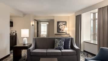 Habitación de hotel con un sofá gris, una mesa de centro, una lámpara, un cuadro en la pared y vistas a la ciudad a través de amplias ventanas.