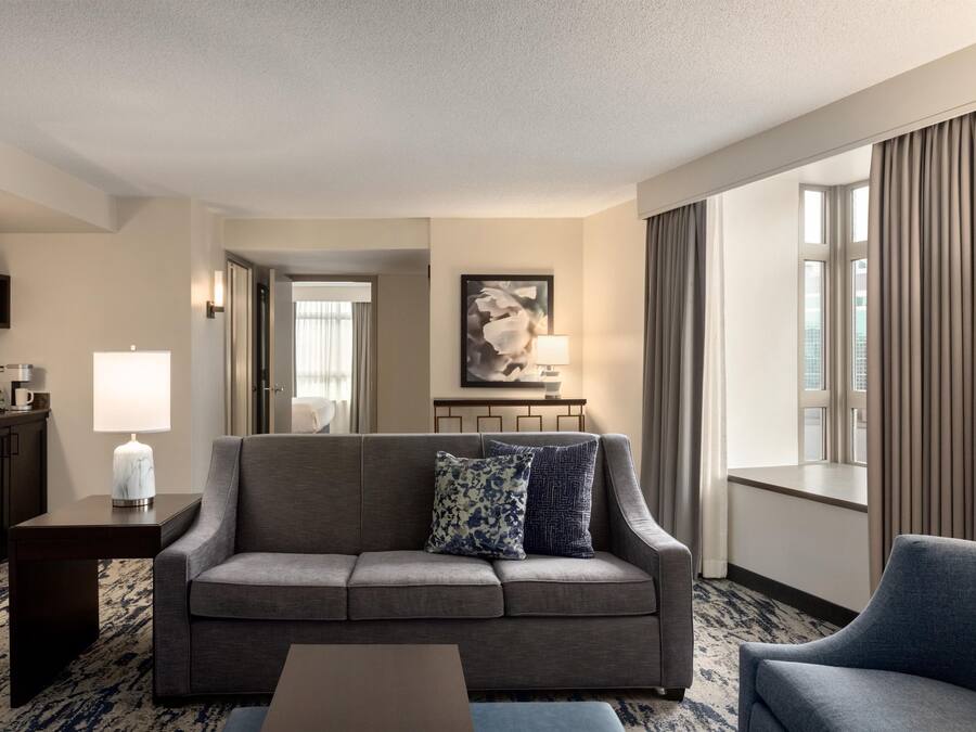 A hotel room with a grey sofa, a coffee table, a lamp, a painting on the wall, and a view of the city through large windows.