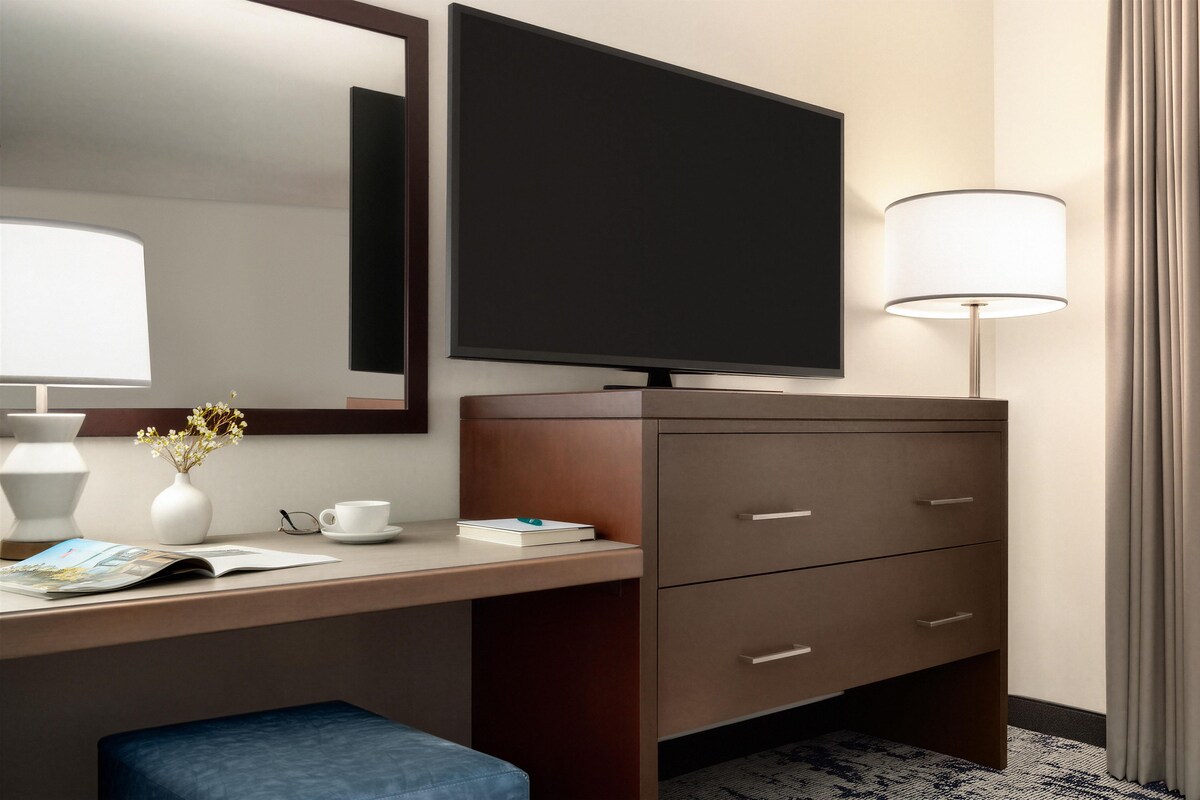 Hotelkamer met flatscreen televisie, bureau met lamp, vaas met bloemen, kop en boek.
