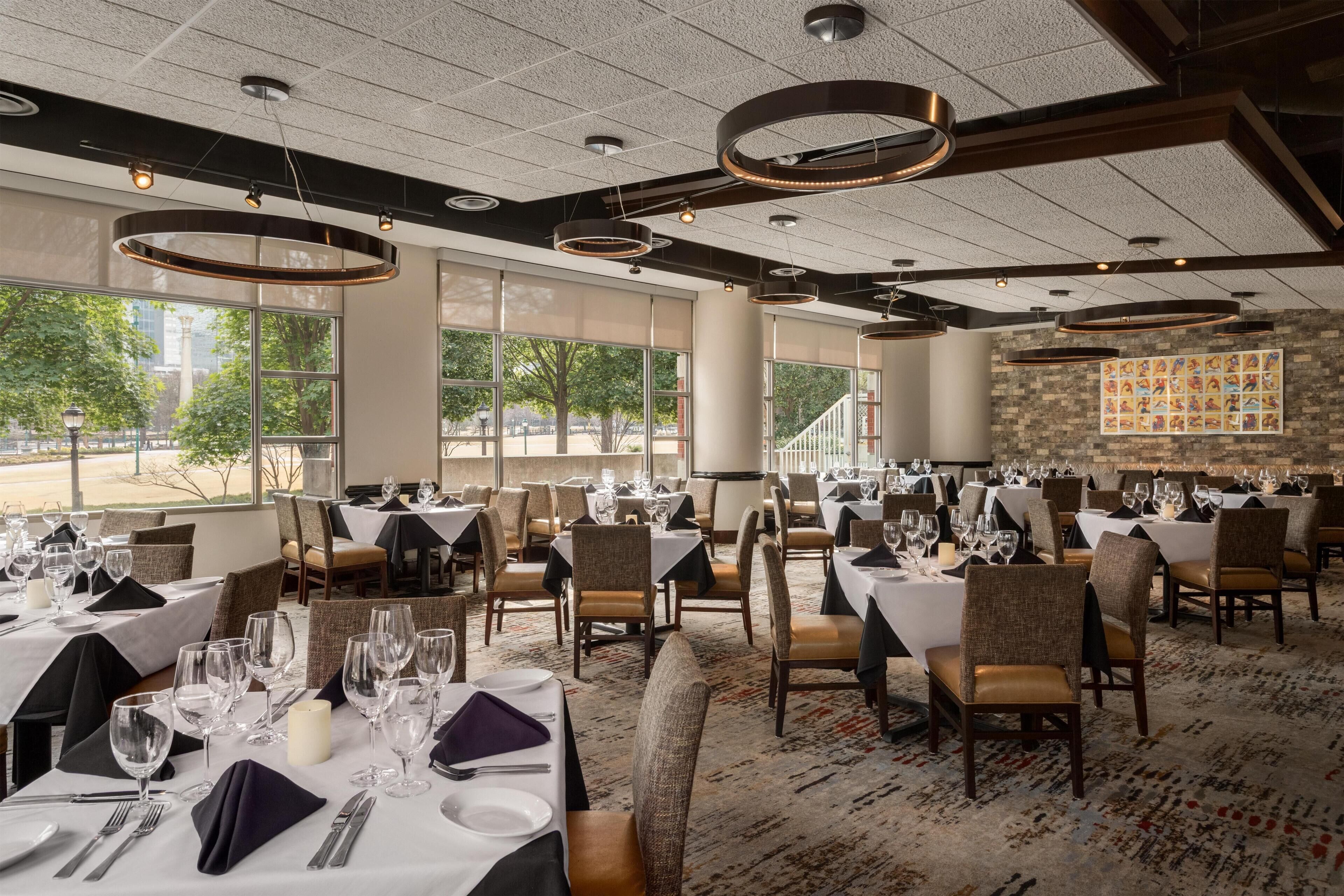 A restaurant with multiple tables set for dining, large windows offering a view of the outdoors, and a modern ceiling design.