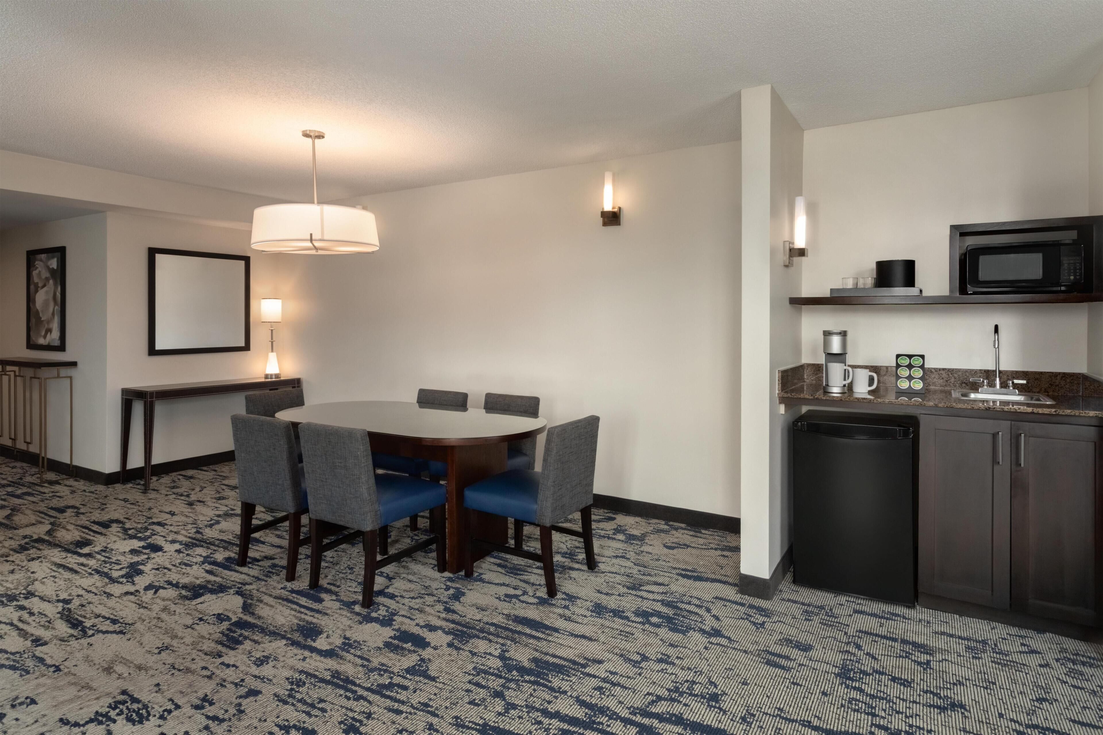 A hotel room with a dining table, chairs, a kitchen area with a microwave, and a coffee maker.