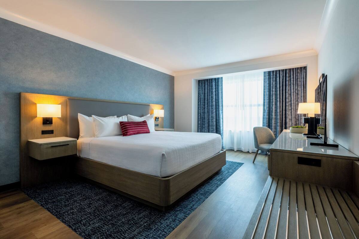 A hotel room with a large bed, bedside lamps, a desk with a lamp, a chair, and a large window with curtains.