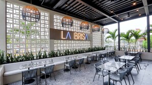 Un espace extérieur aménagé avec des tables et des chaises, un mur décoratif orné de l’enseigne « LA BRISA », et une vue sur des palmiers.