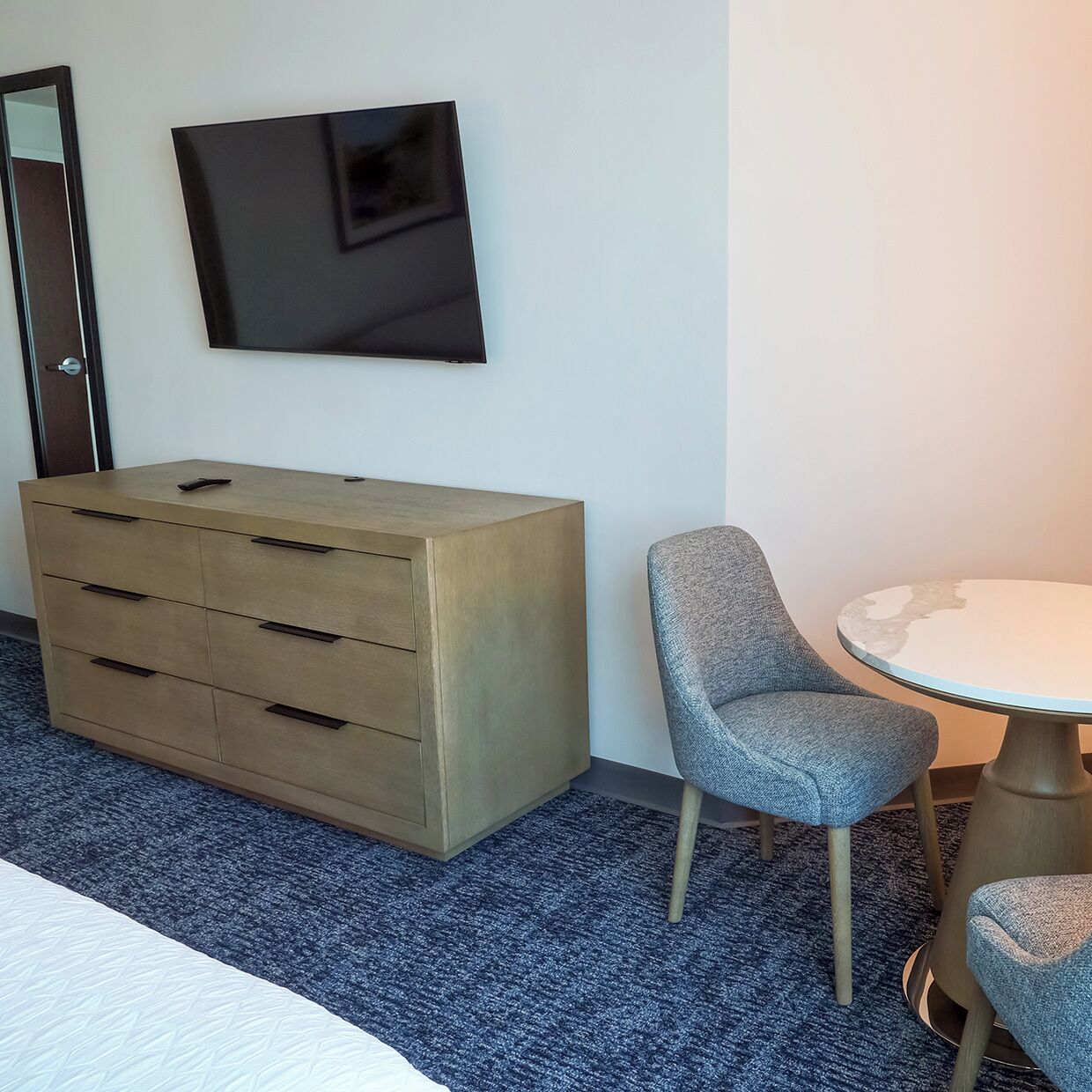 A hotel room with a wooden dresser, a gray chair, a small round table, and a flat-screen TV mounted on the wall.
