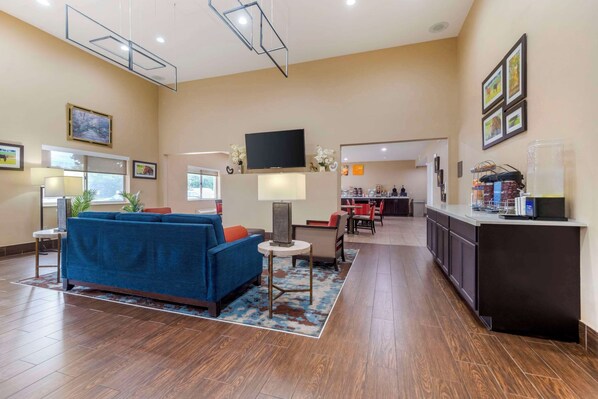 A hotel lobby with a blue sofa, a small table, a TV, and a kitchen area in the background.