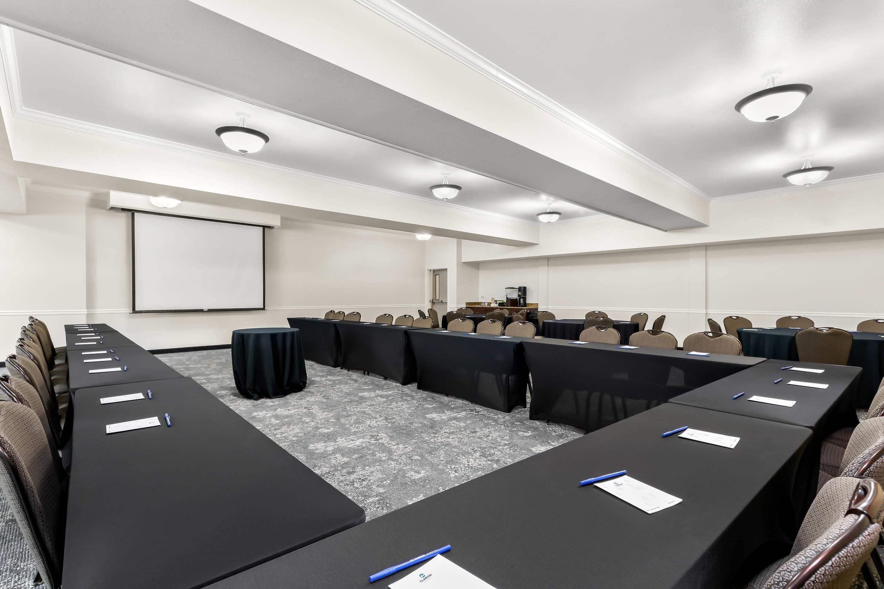 A conference room with long tables, chairs, a projector screen, and a coffee station.