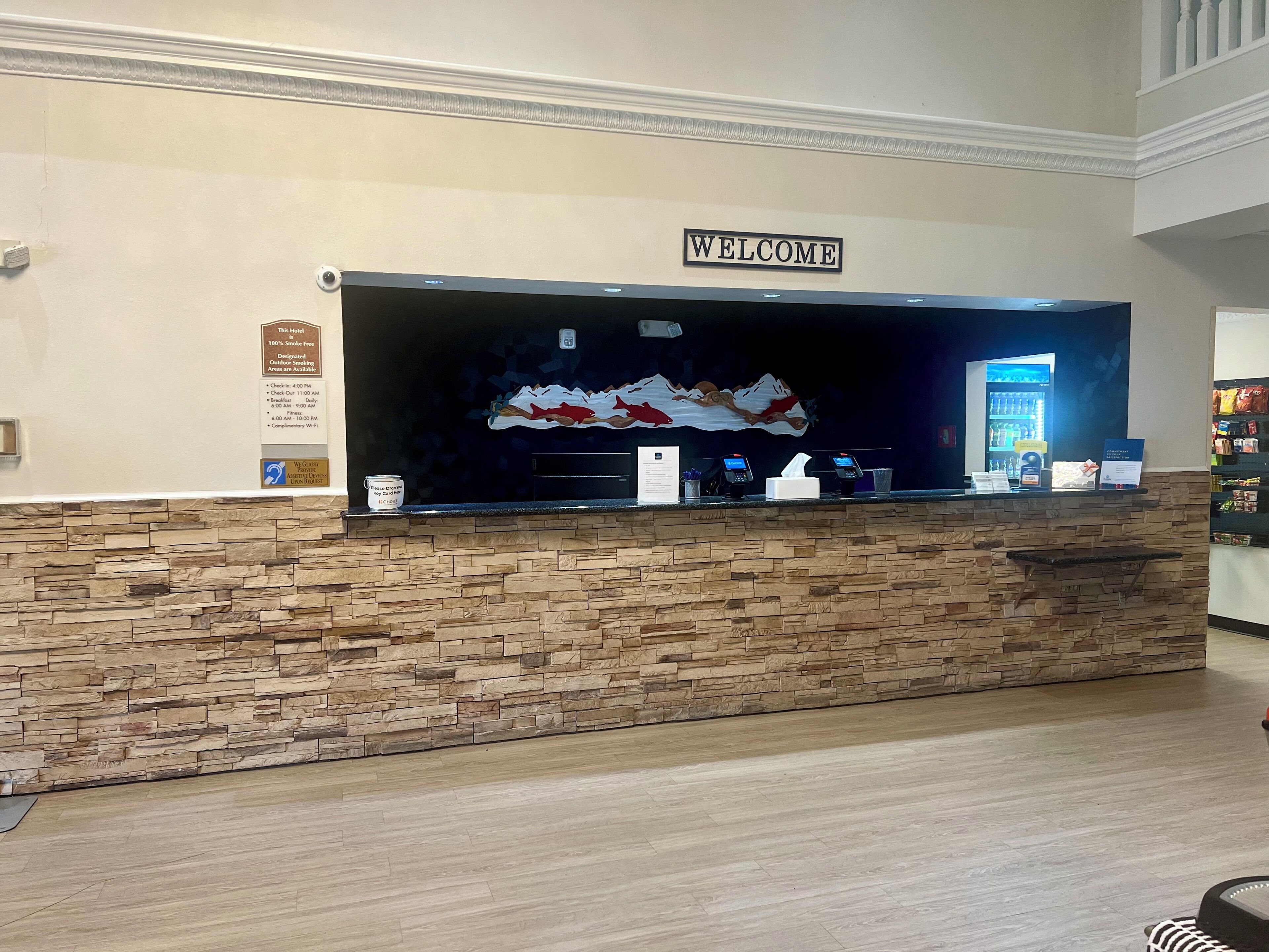 A hotel reception desk with a 'WELCOME' sign, a stone wall, a digital display, and a sign indicating the hotel is smoke-free.
