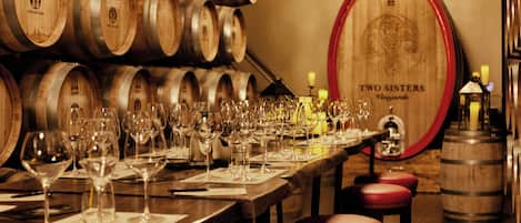 Una sala de cata de vinos con barricas de madera, una gran barrica de madera con la etiqueta 'Two Sisters' y varios vasos de vino sobre una mesa larga.