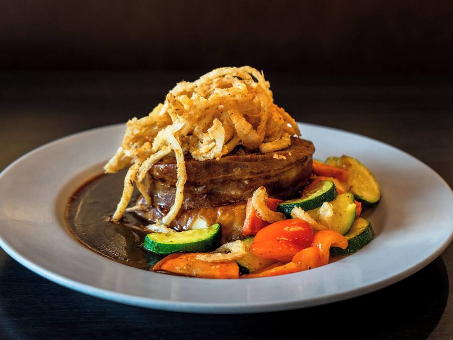 A plate of food with a steak topped with fried onions, accompanied by sliced zucchini, carrots, and bell peppers.