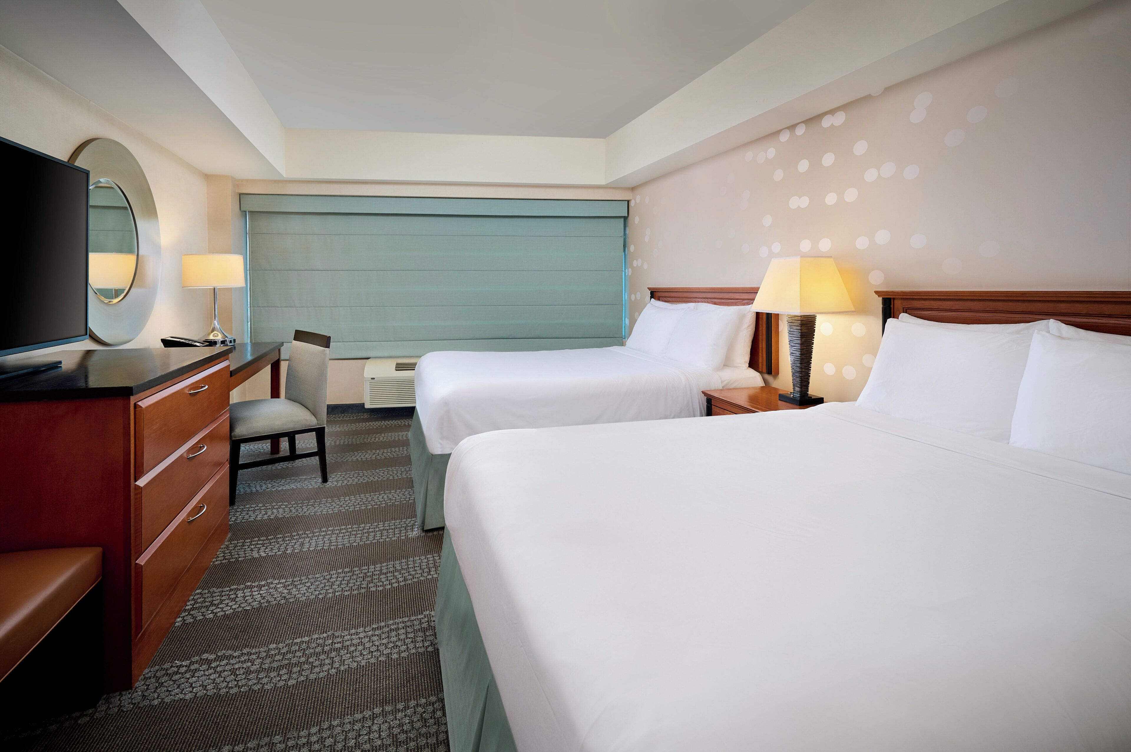 A hotel room with two beds, a desk with a chair, a television, and a wall-mounted lamp.