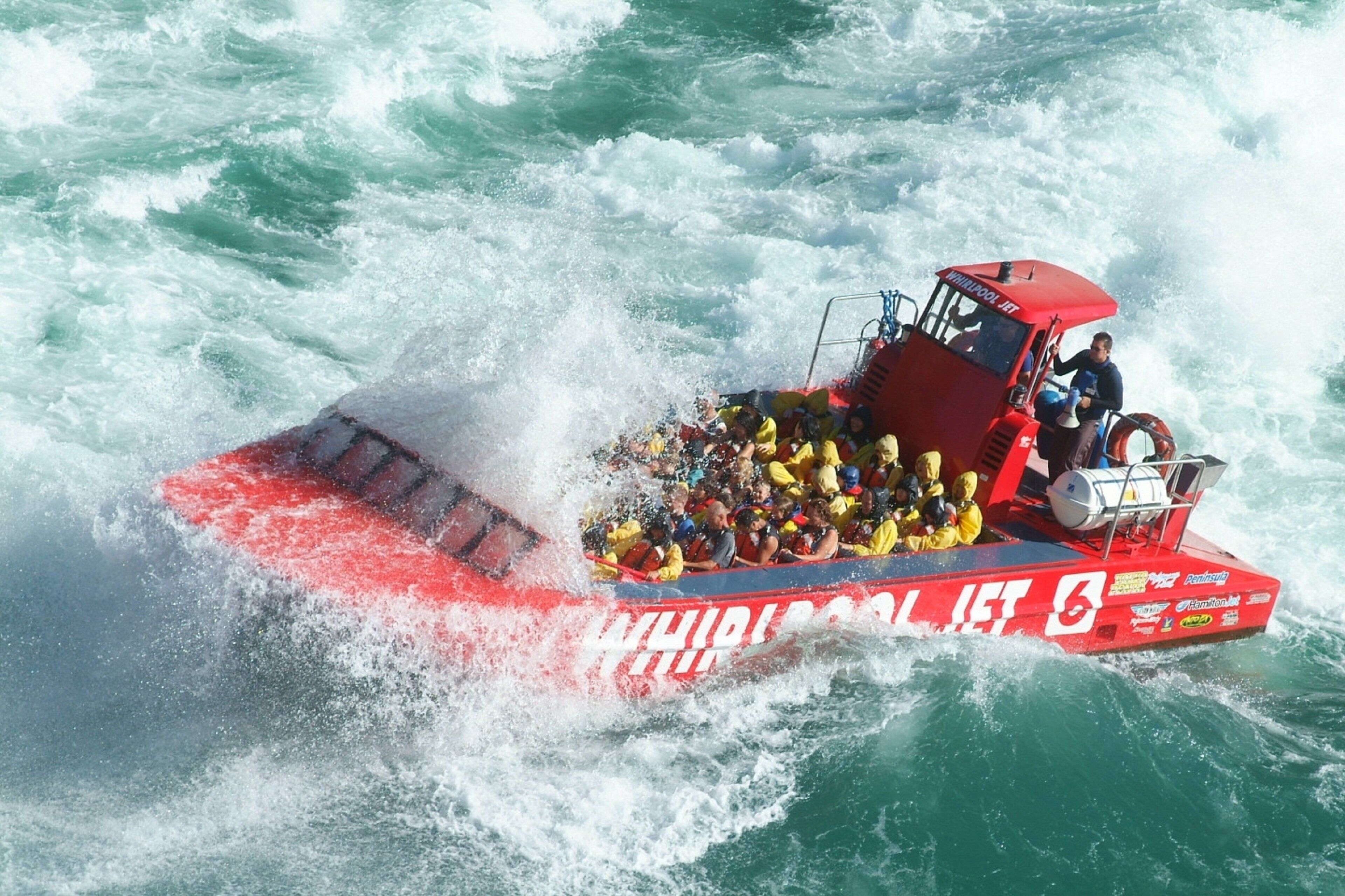 Un bateau à moteur rouge, sur lequel on peut lire les inscriptions « WHIRLPOOL JET » et « 5 », est rempli de personnes portant des gilets de sauvetage jaunes et navigue sur des eaux agitées.