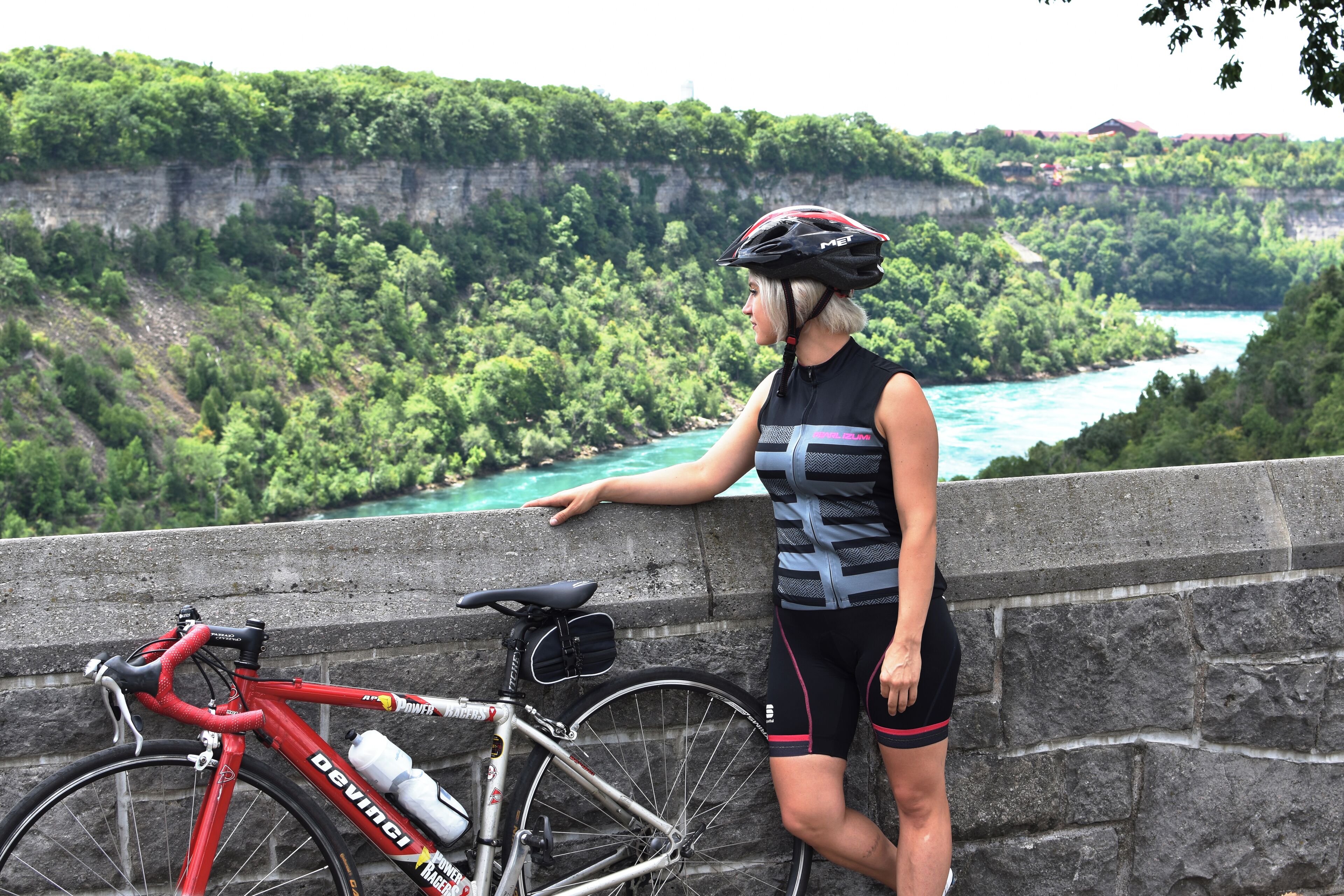A person leaning on a stone wall overlooking a river and cliffs, with a red bicycle and water bottle beside them.