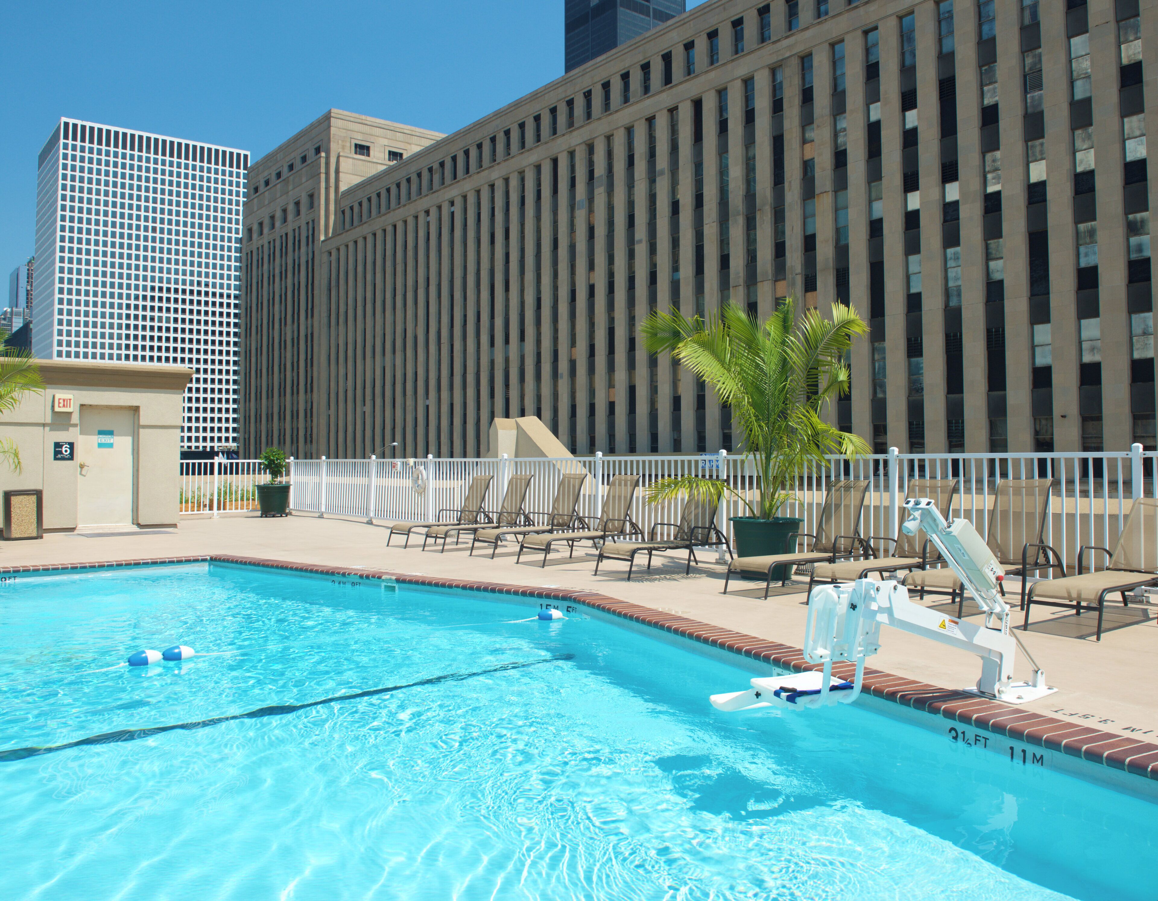 A swimming pool with lounge chairs and a lifeguard chair, surrounded by a fence, with tall buildings in the background.