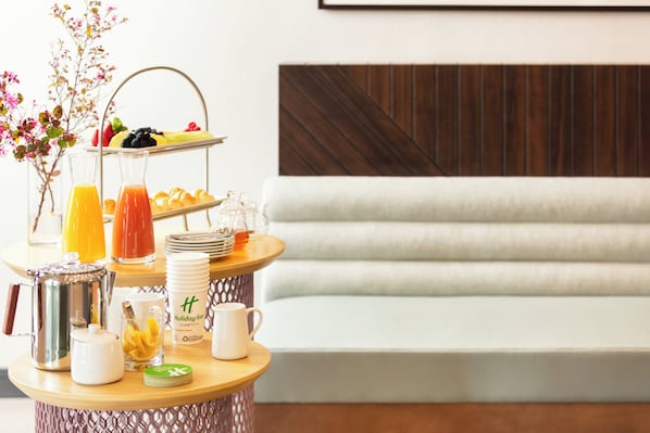 Un bufé de desayuno con una variedad de frutas, pasteles y bebidas sobre una mesa de madera, en un espacio moderno con un sofá blanco y un panel de madera decorativo.