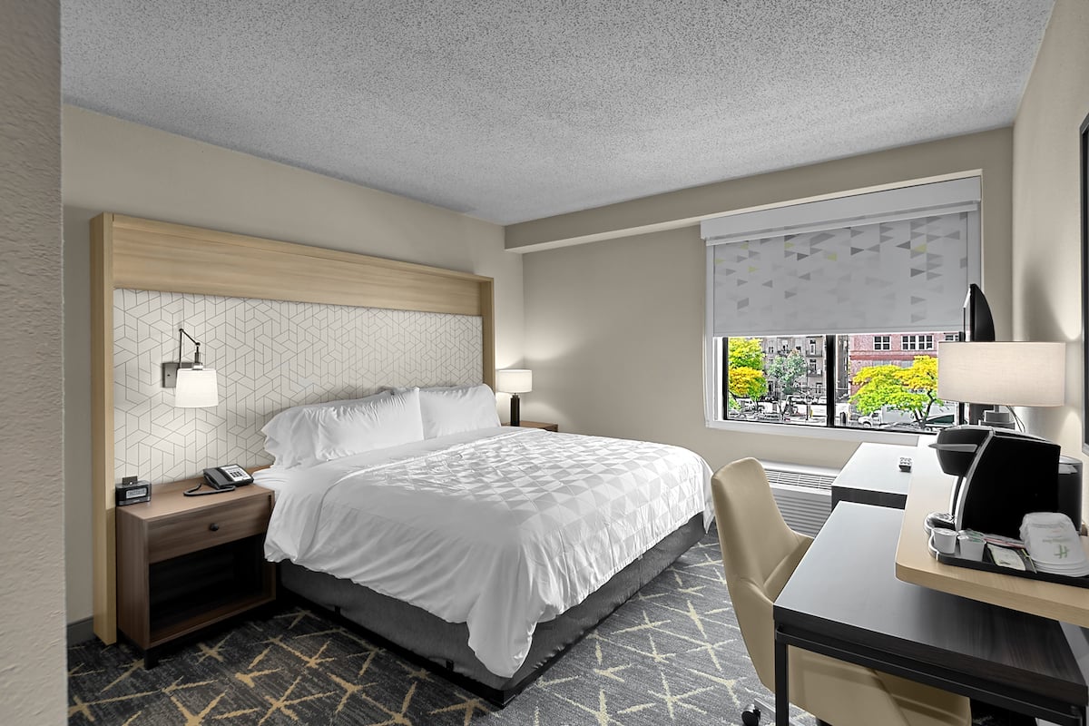 A hotel room with a bed, bedside tables, a desk, and a window with a view of the city.
