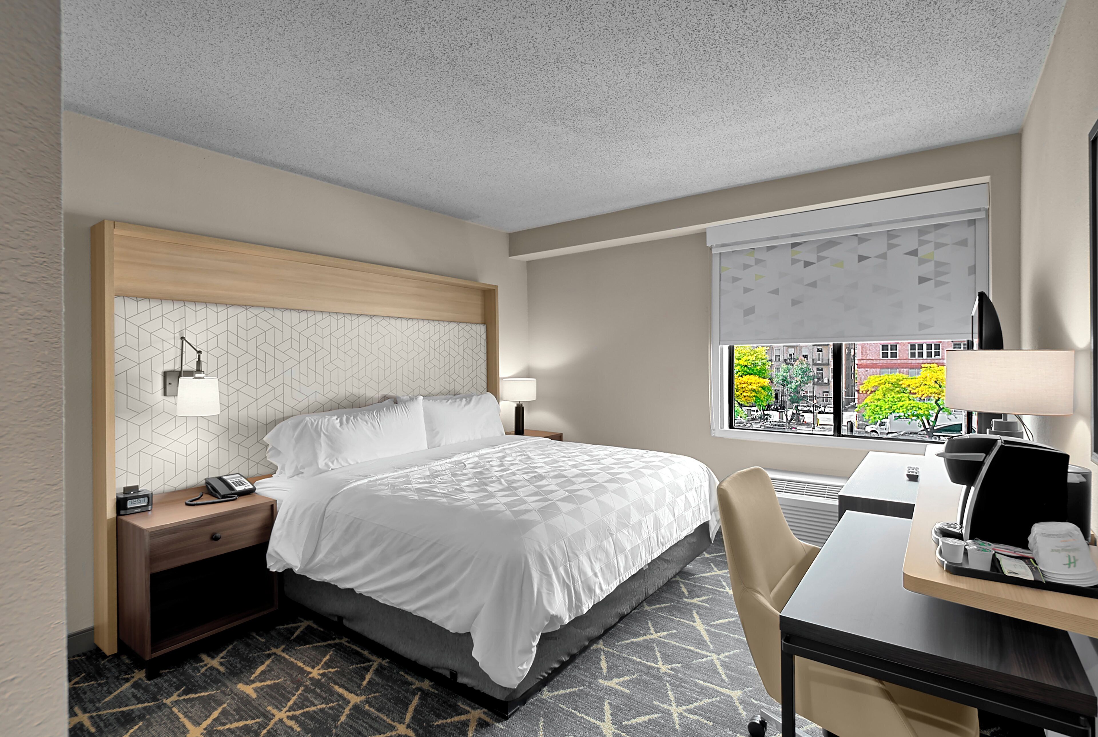 Une chambre d’hôtel comprenant un lit, une table de chevet, un bureau, une chaise et une fenêtre donnant sur des bâtiments et des arbres.