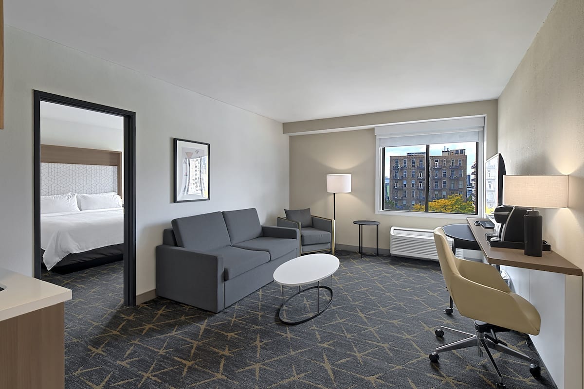 A hotel room with a bed, a gray sofa, a small white table, a desk with a chair, and a window with a city view.