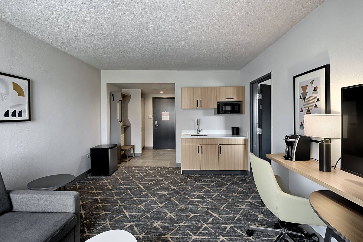 A modern hotel room with a kitchenette, a grey sofa, a small table, and a patterned carpet.