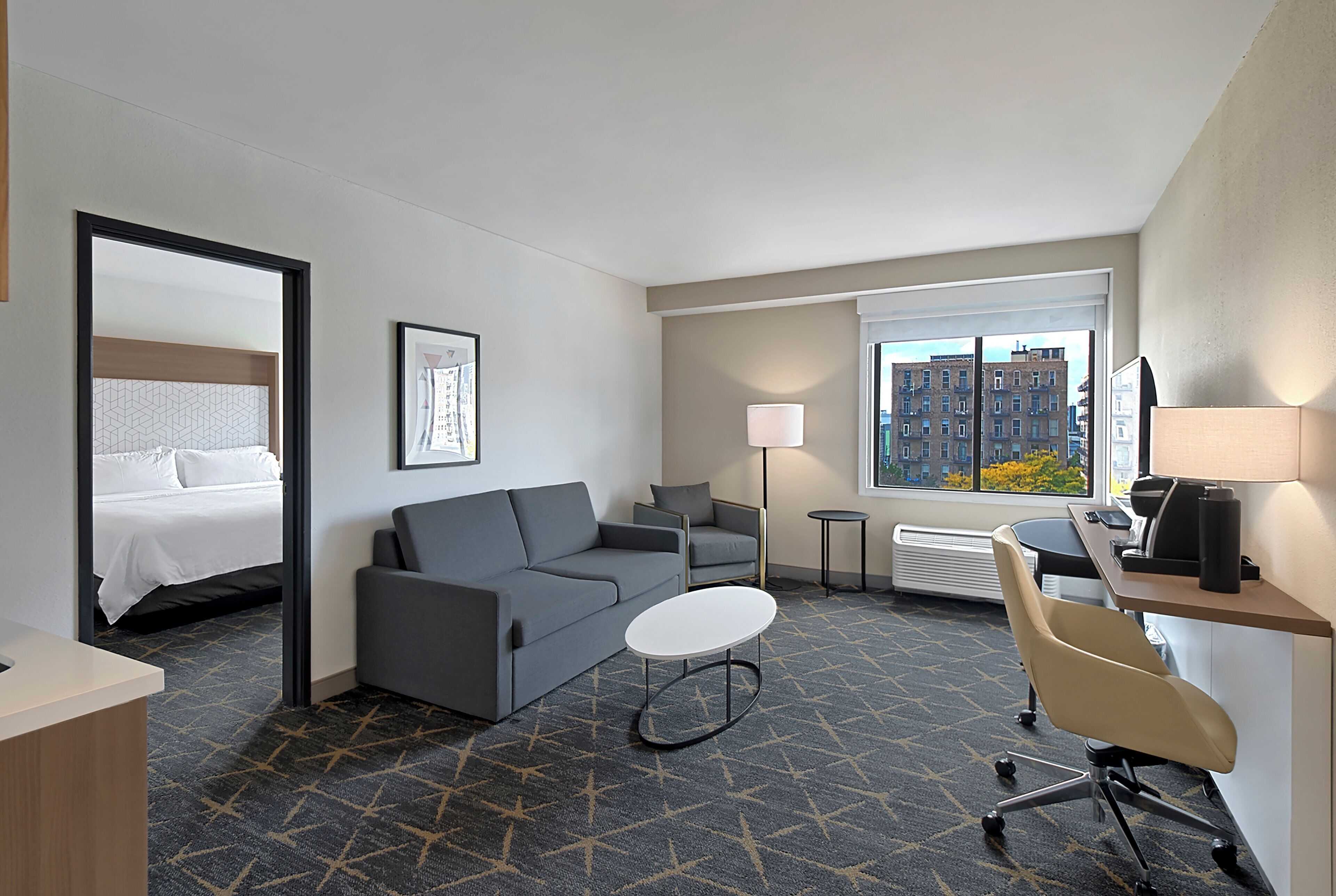 A hotel room with a bed, a grey sofa, a small round table, a desk with a chair, a standing lamp, and a window with a city view.