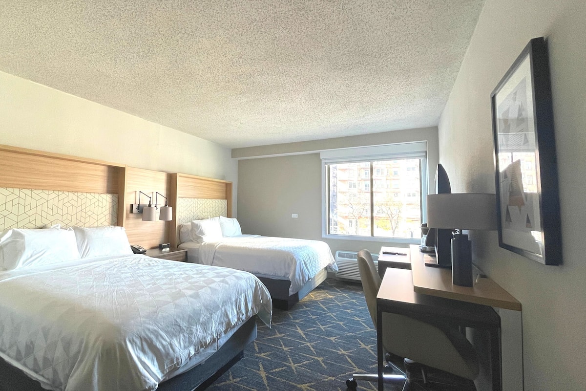 A hotel room with two beds, a desk, a chair, a TV, and a window with a view of buildings.