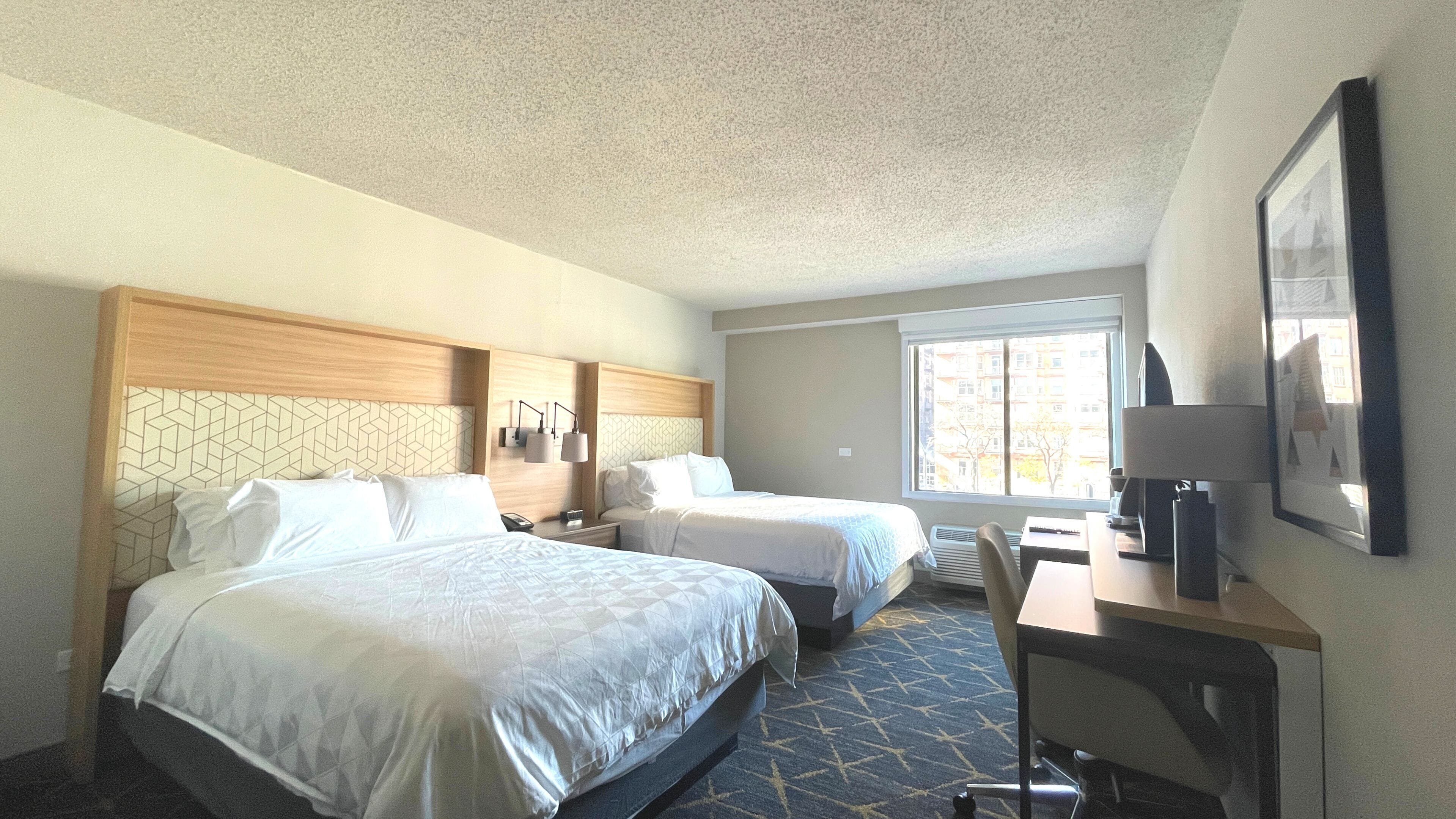 A hotel room with two beds, a desk, a chair, a TV, and a window with a view of buildings.