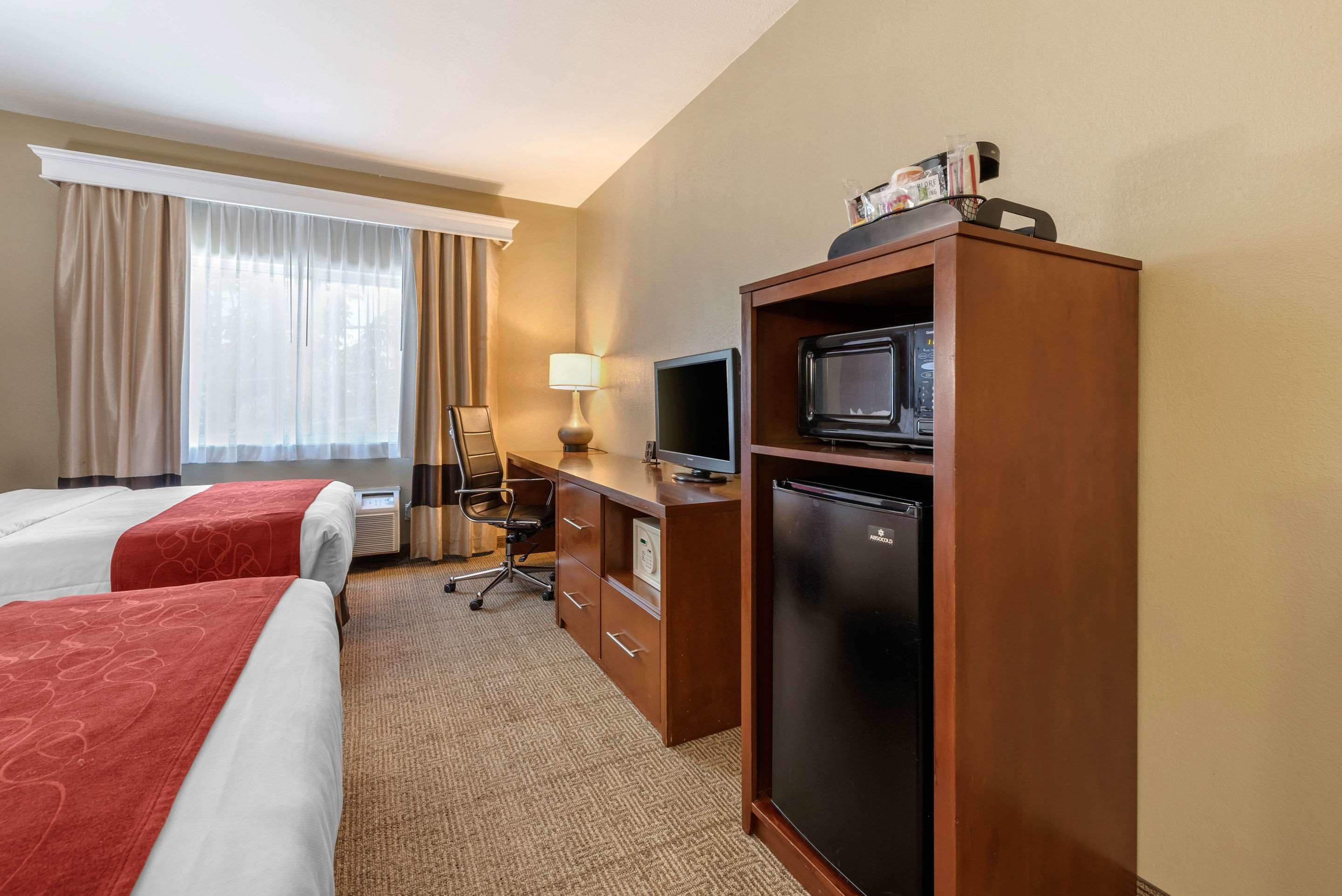 A hotel room with two beds, a desk with a computer and a microwave, a refrigerator, and a window with curtains.