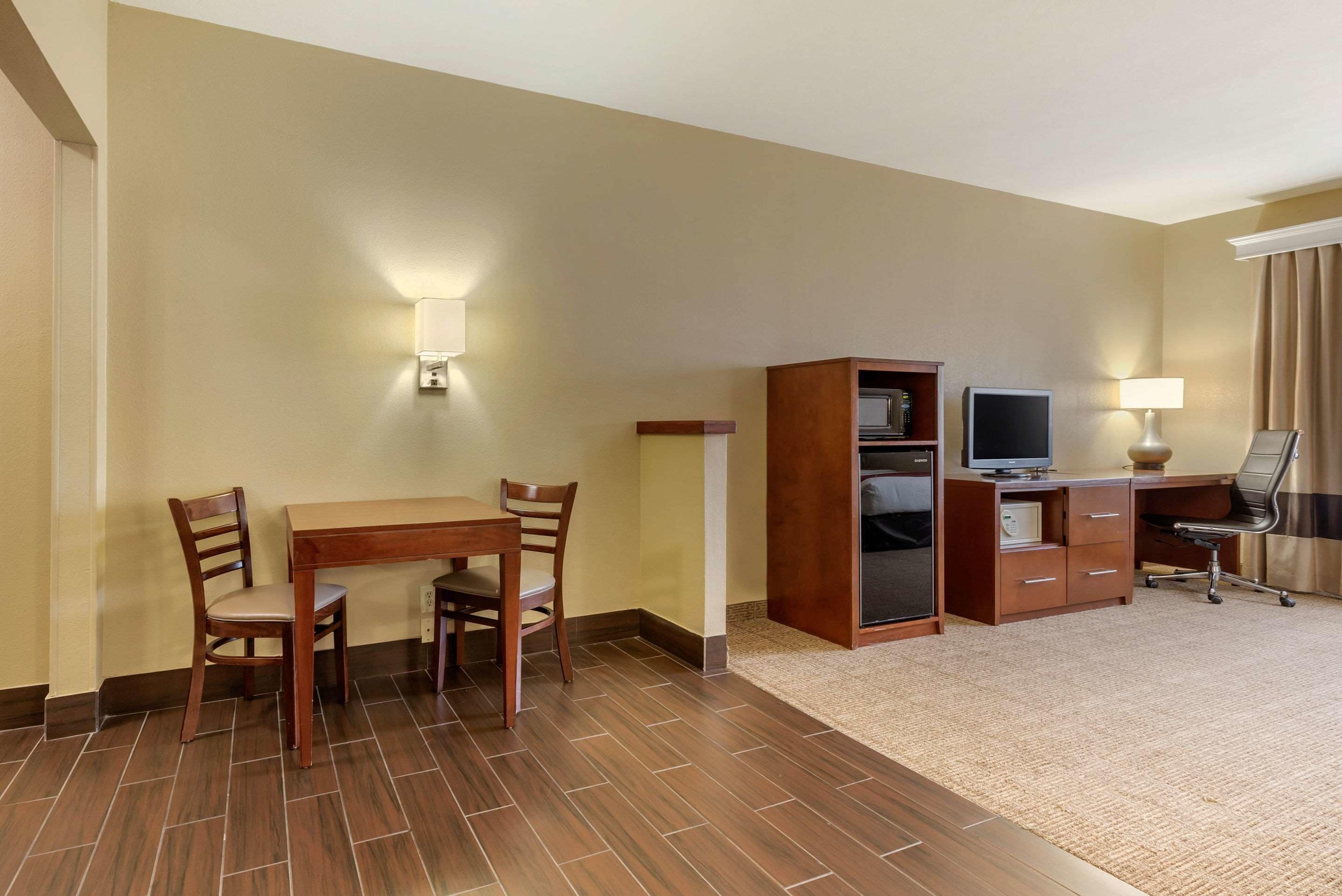 A hotel room with a wooden dining table, two chairs, a TV, a desk, and a lamp.
