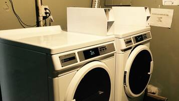 A front-loading washer and dryer unit with a digital display, located in a laundry room with visible instructions and a sign indicating guest laundry hours.