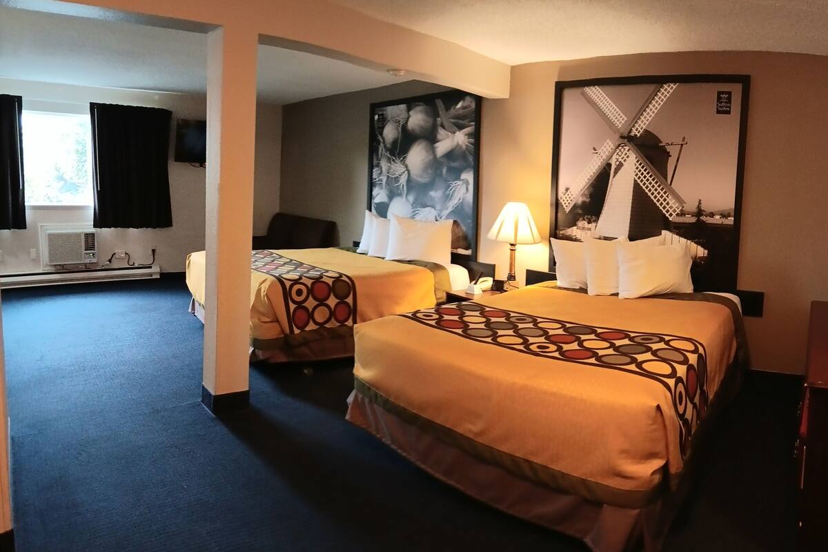 Habitación de hotel con dos camas, un escritorio, una silla, una lámpara y un ventanal grande con cortinas.