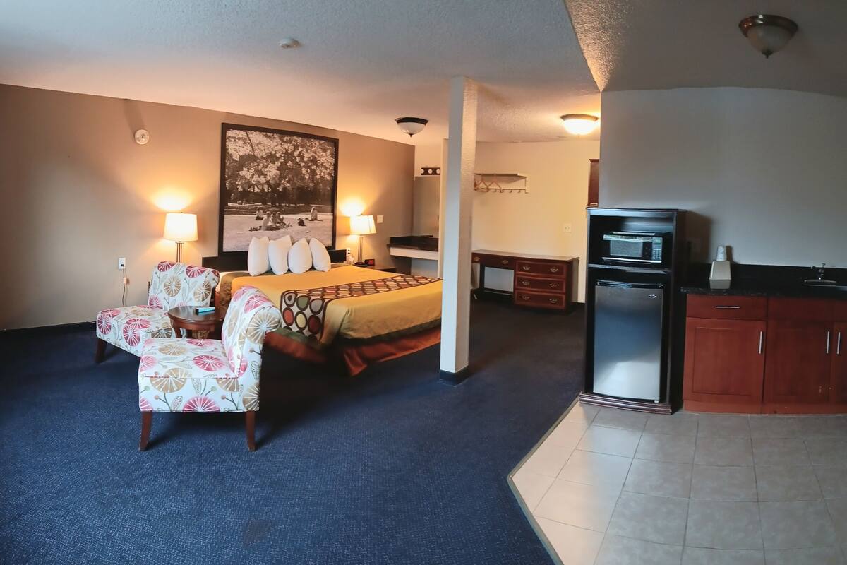 Habitación de hotel con cama, dos sillas, escritorio, refrigerador y un cuadro en la pared.