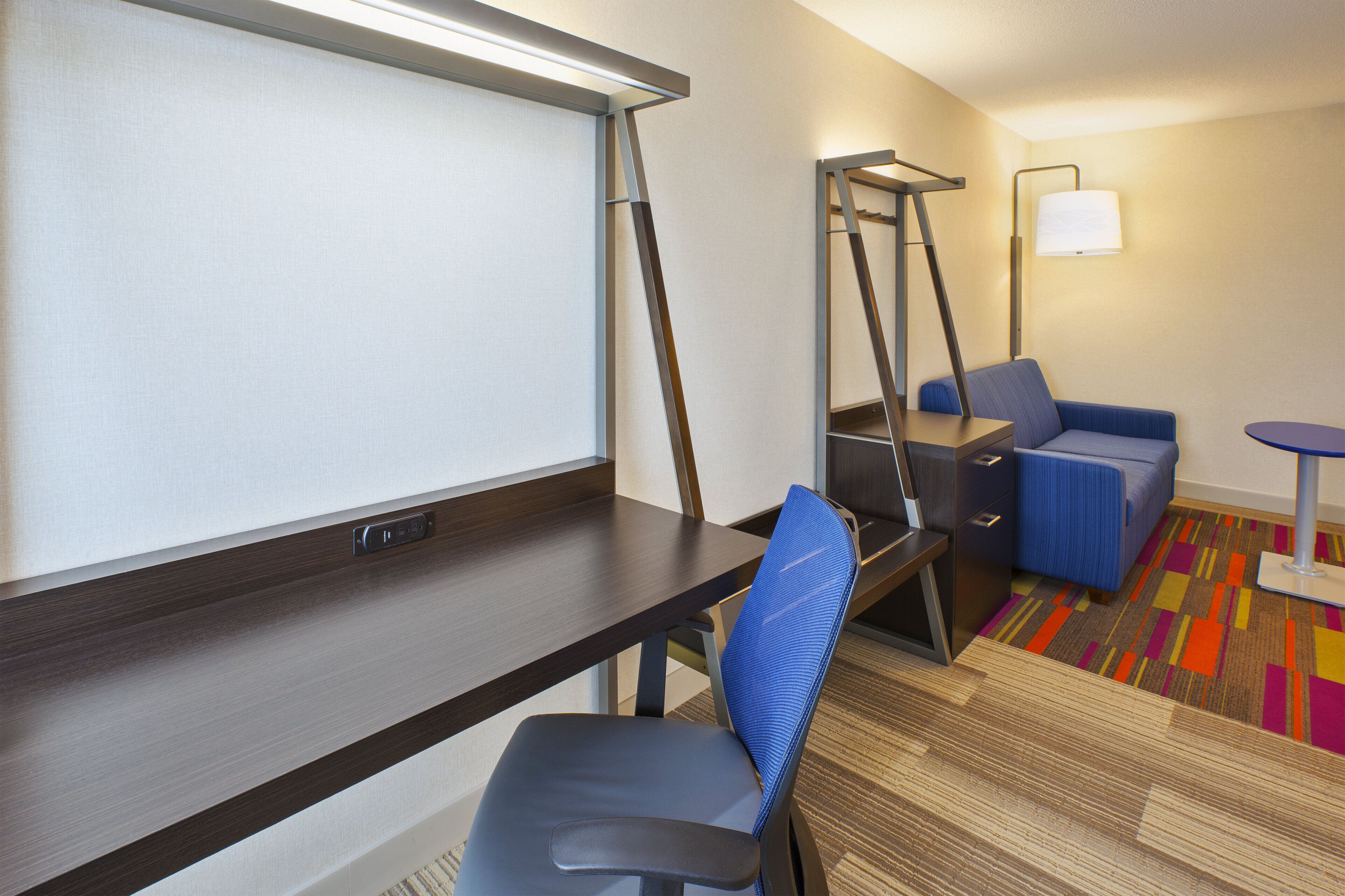 A hotel room with a desk, a chair, a bed, a small table, a lamp, and a colorful carpet.