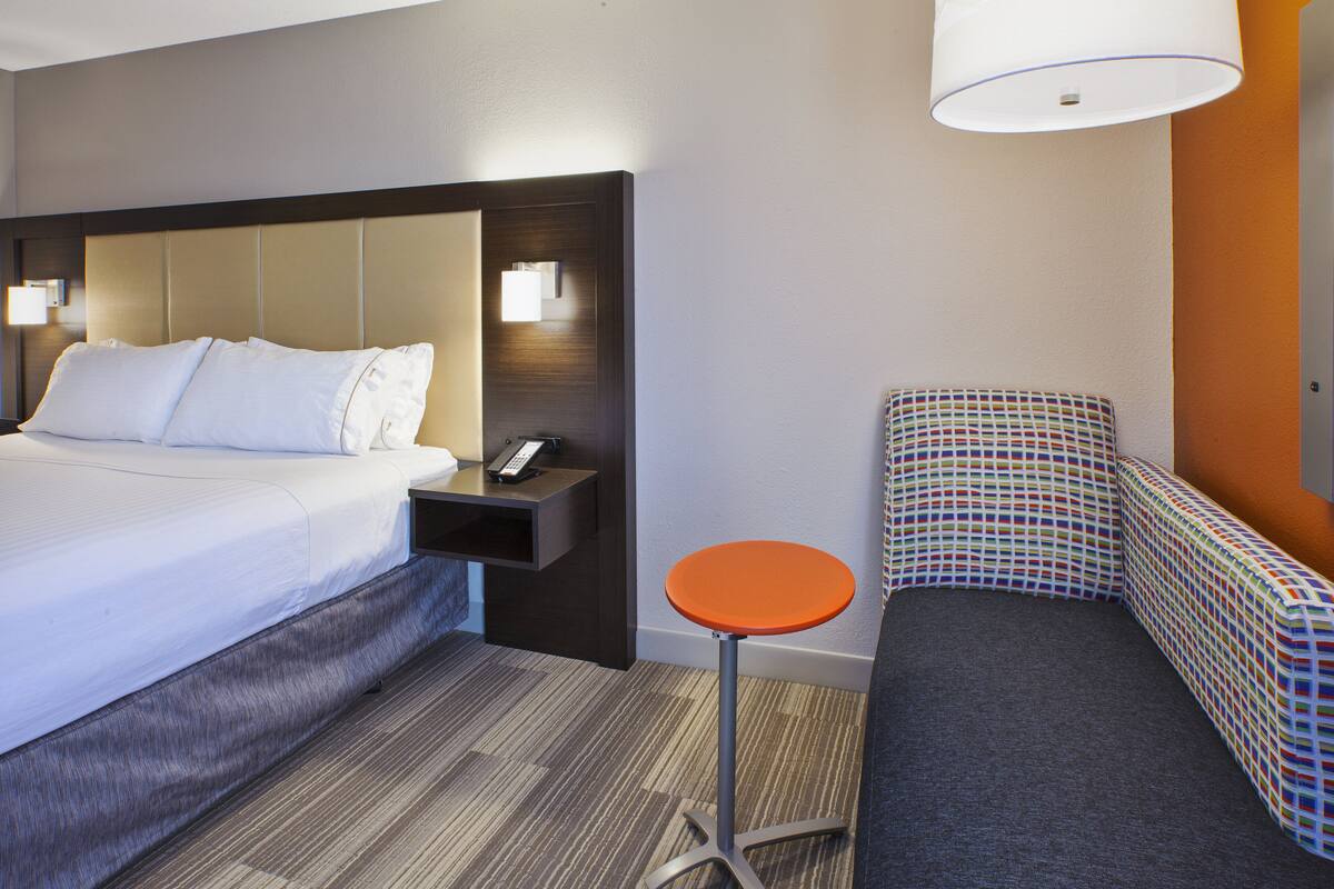A hotel room with a bed, bedside table, lamp, and a small stool.