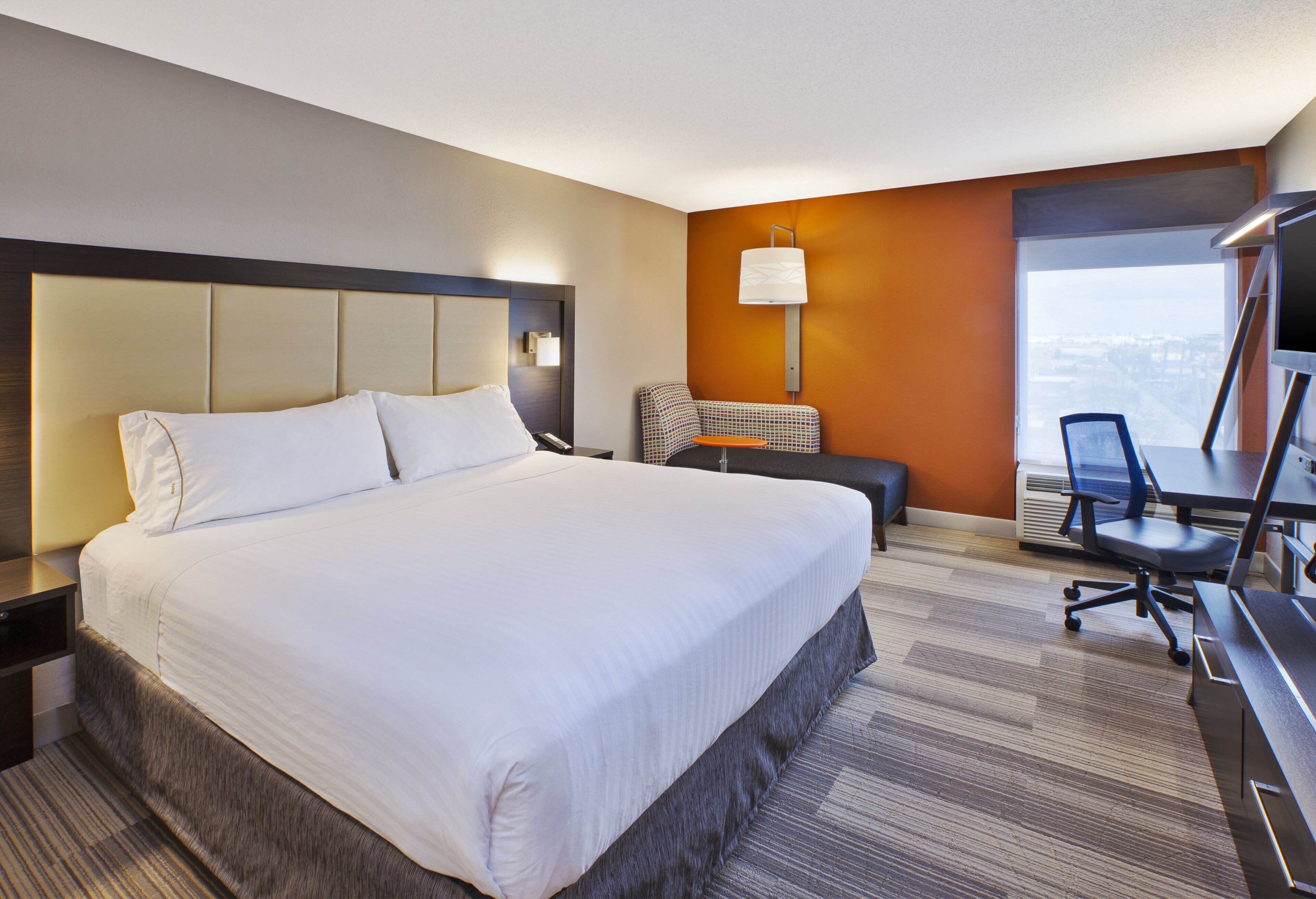 A hotel room with a large bed, a desk with a chair, a lamp, and a view of the city.