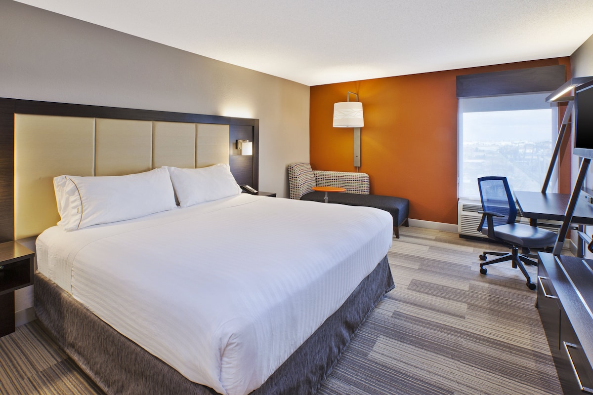 A hotel room with a large bed, a desk with a chair, a lamp, and a view of the city.
