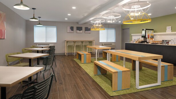 A modern dining area with wooden tables and benches, green chairs, and pendant lighting.