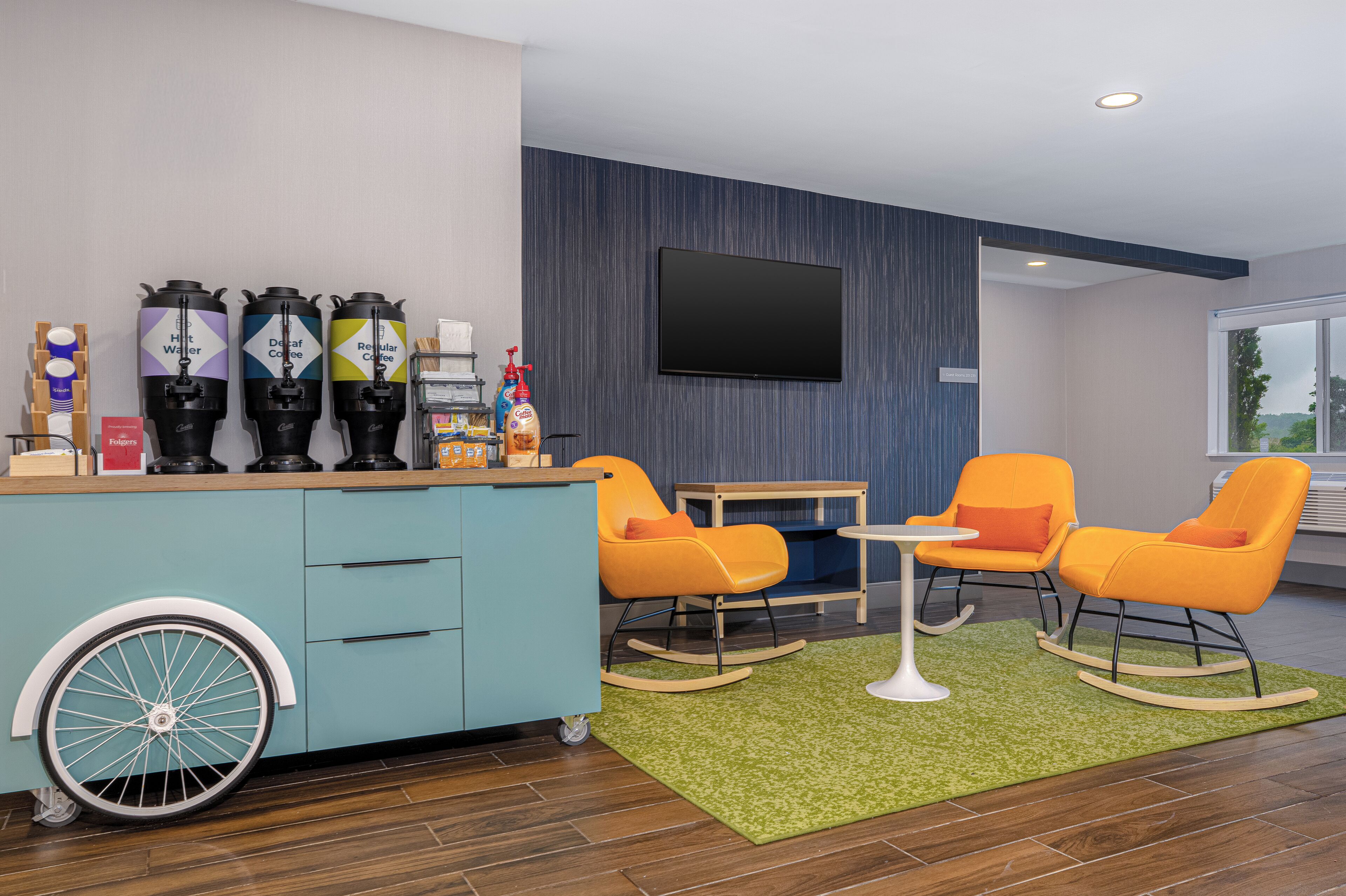 A modern room with a blue wall, a mounted TV, a coffee station with dispensers, and orange rocking chairs.