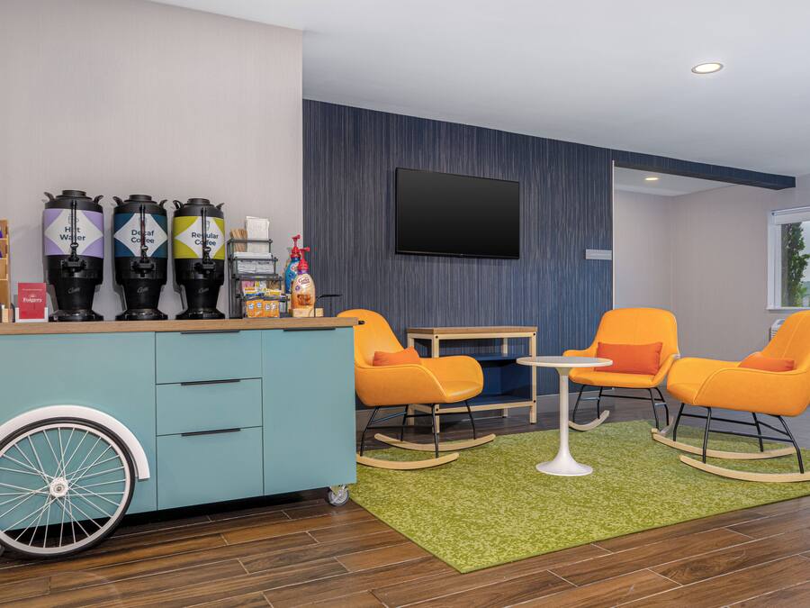 A modern room with a blue wall, a mounted TV, a coffee station with dispensers, and orange rocking chairs.