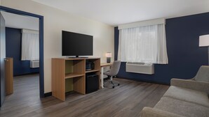 A hotel room with a wooden desk, a television, a grey office chair, a sofa, and a window with curtains.