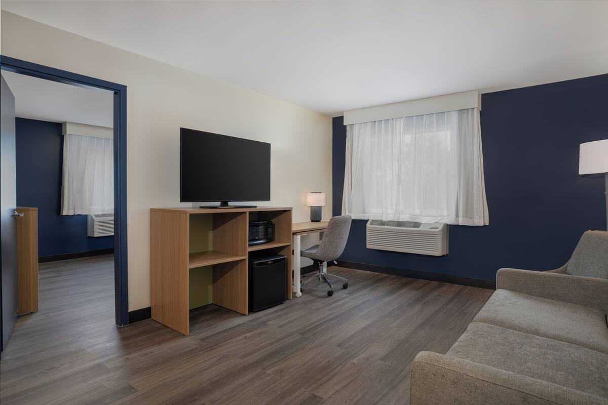 A hotel room with a wooden desk, a television, a grey office chair, a sofa, and a window with curtains.