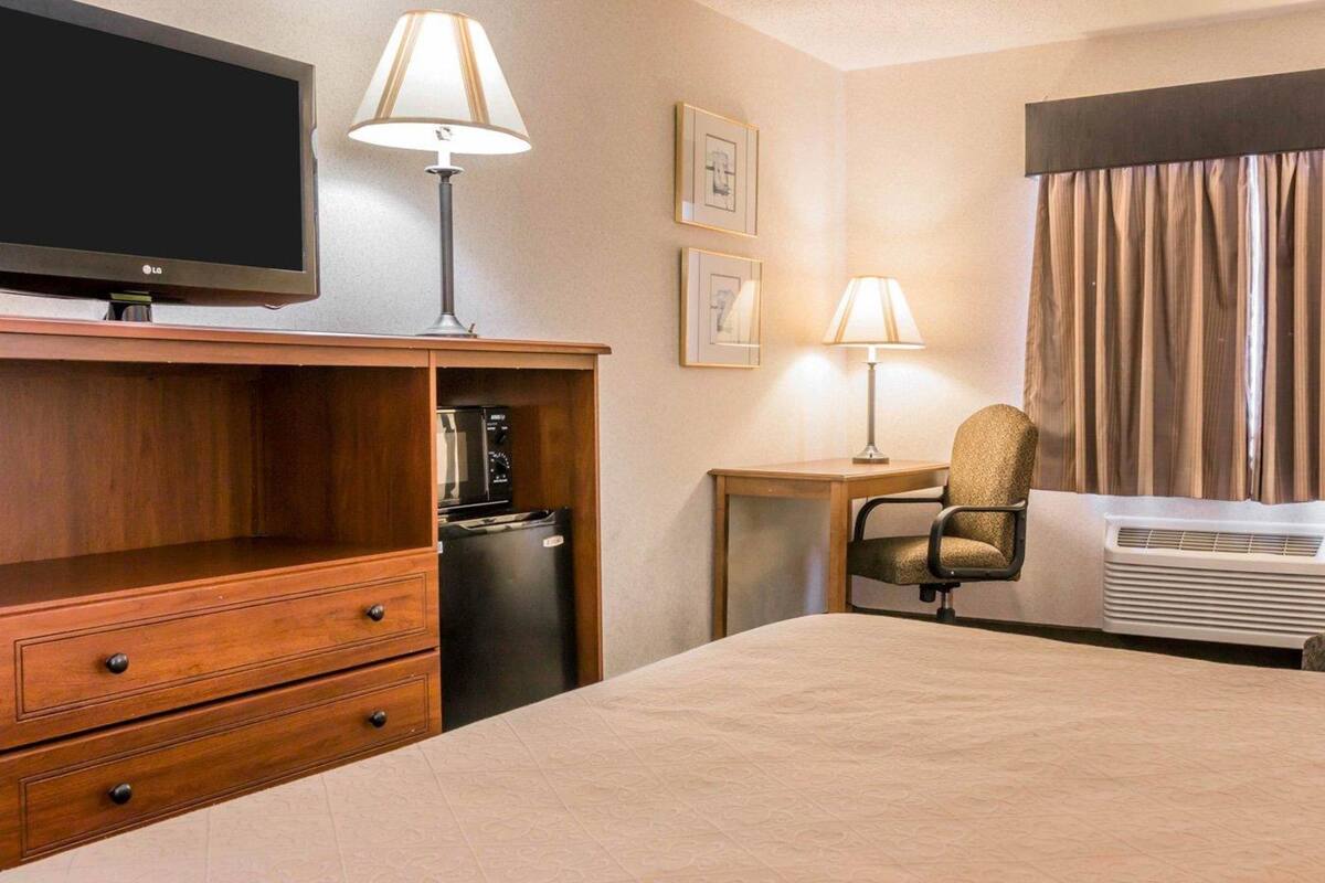 A hotel room with a bed, a TV, a desk with a lamp, a chair, a refrigerator, and an air conditioner.