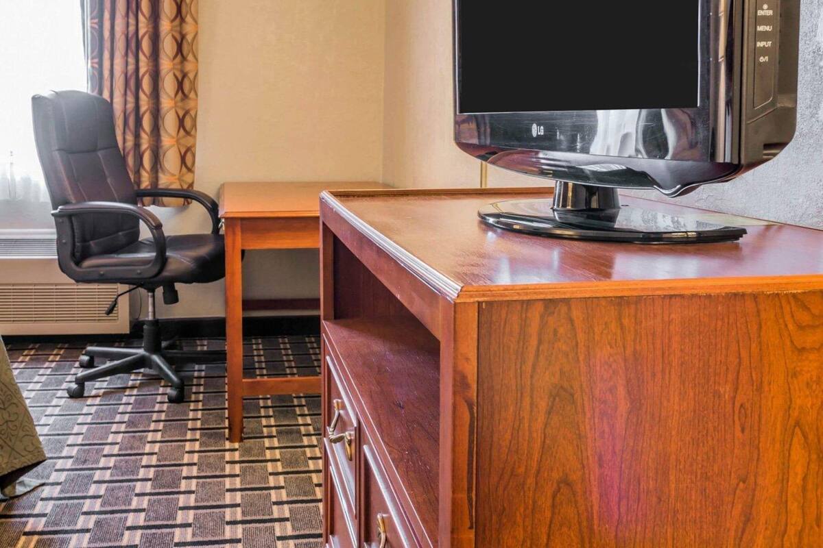 A hotel room with a wooden desk, a black office chair, a television, and a window with patterned curtains.