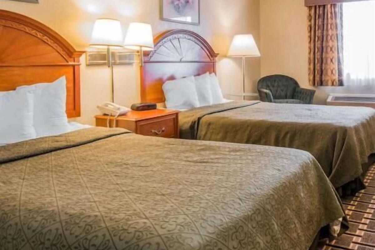A hotel room with two beds, a wooden headboard, a nightstand with a lamp, a clock, and a chair.