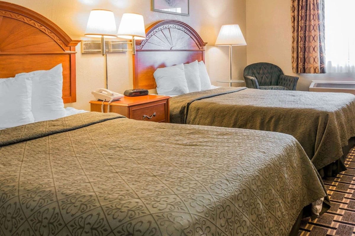 A hotel room with two beds, a wooden headboard, a nightstand with a lamp, a chair, and a window with curtains.