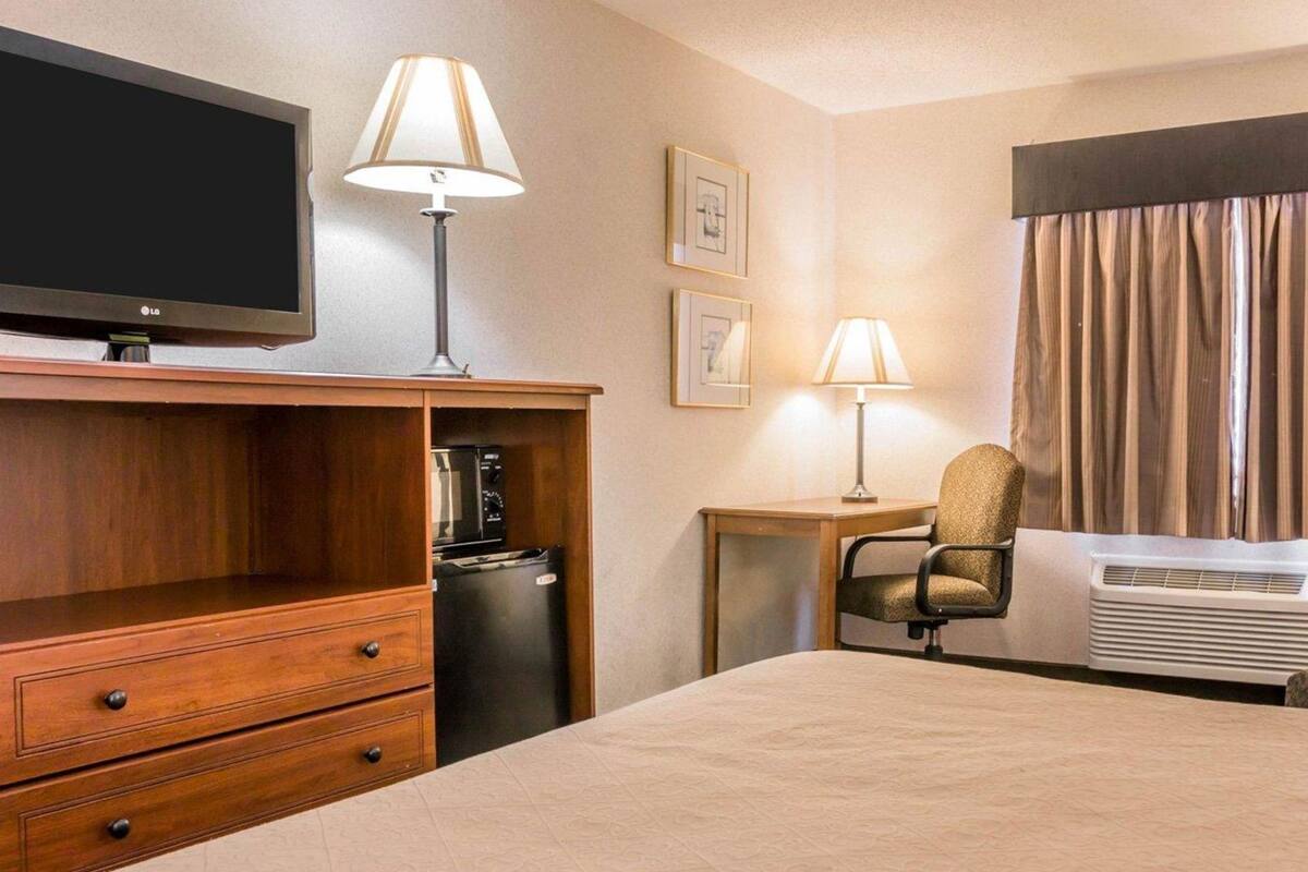 A hotel room with a bed, a TV, a desk with a lamp, a chair, a refrigerator, and a window with curtains.