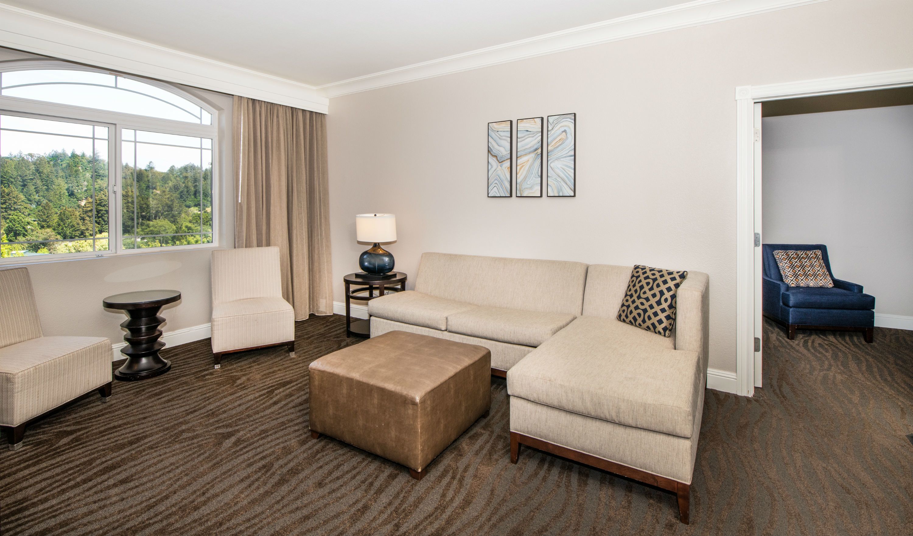 A hotel room with a large window, a beige sofa, a matching armchair, a small round table with a lamp, and a blue armchair with a patterned pillow.
